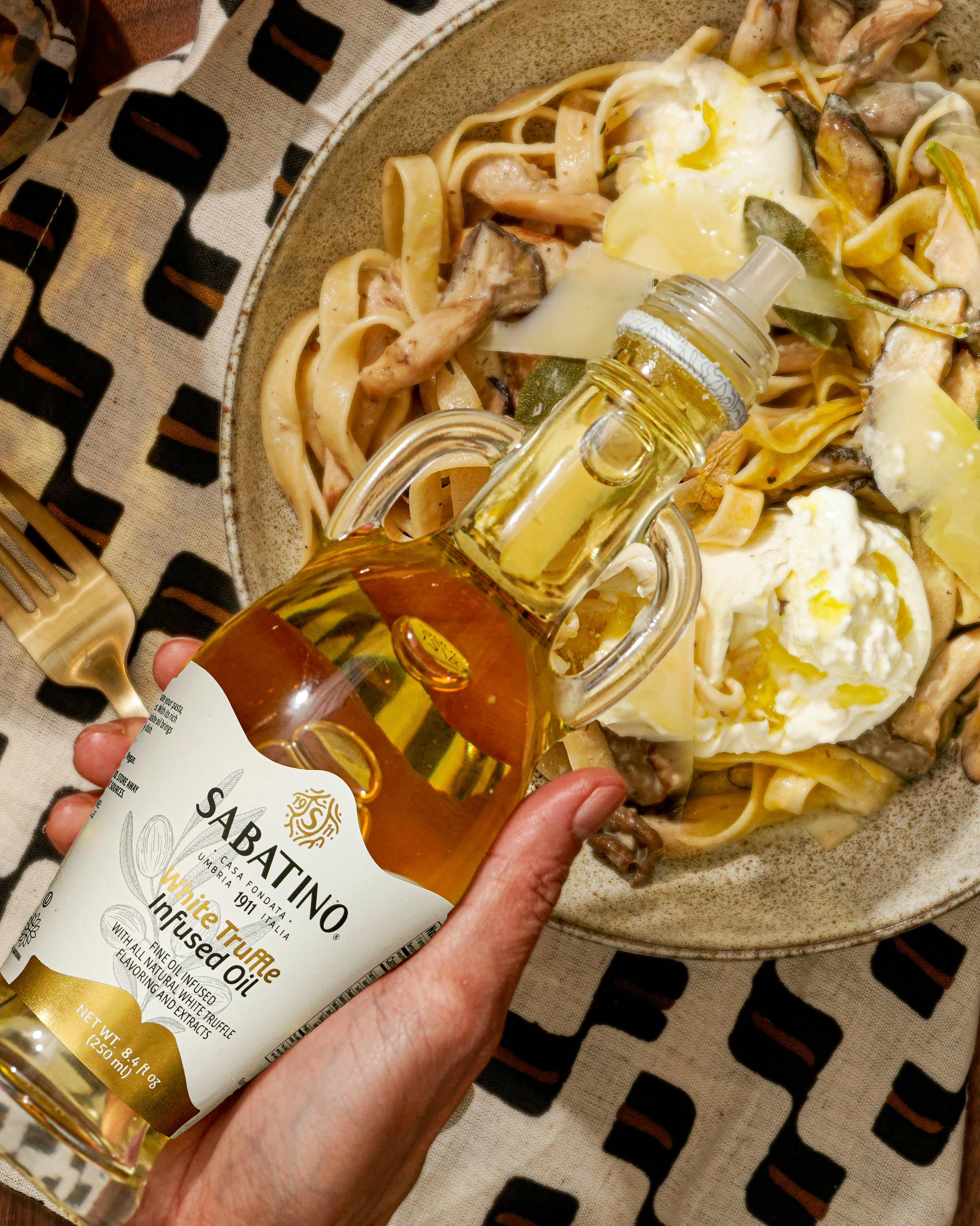 a hand holding a bottle of oil next to a bowl of pasta